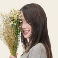 ストレートの黒髪の人が白と黄色の小さな花のブーケを持ち、顔の片側を部分的に覆い、カメラに向かって微笑んでいるIcons8 Teamの写真。背景は無地で明るい色です。.